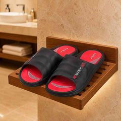 A pair of black slide sandals with bright red footbeds and white branding, displayed on a wooden wall-mounted shelf in a bathroom setting.